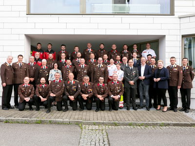 Gruppenfoto von Einsatzkräften und Feuerwehrleuten vor einem modernen Gebäude.